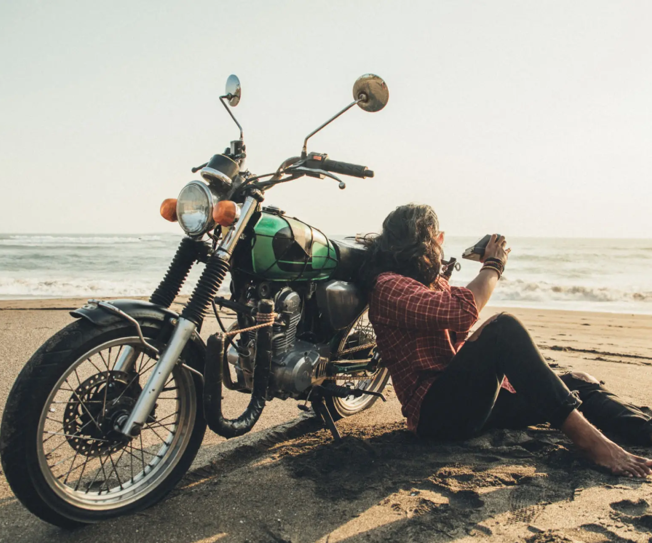 Person relaxing by a motorcycle on a beach at sunset.