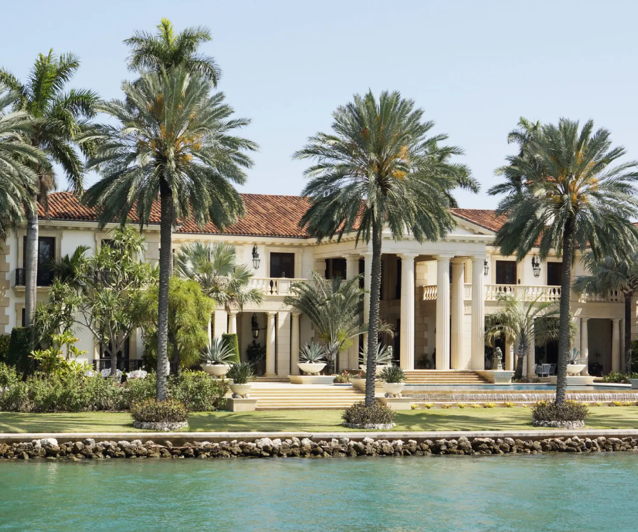 Luxurious waterfront mansion with palm trees and classical architecture.